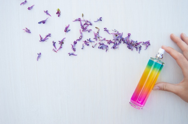 Mão ao lado de pétalas roxas e frasco de perfume multicolorido em um fundo branco de madeira