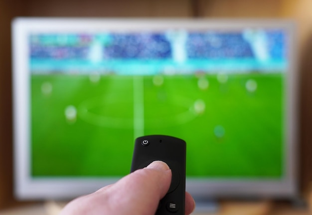 mão apontando o controle remoto no aparelho de tv com futebol ou partida de futebol