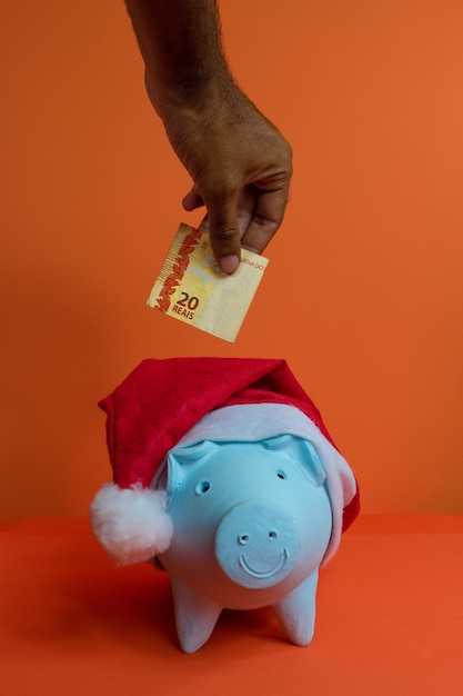 Mão colocando dinheiro brasileiro real no cofrinho com chapéu de Papai Noel de Natal isolado em fundo laranja. Imagem do conceito.