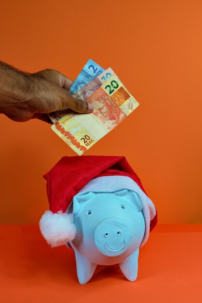 Mão colocando dinheiro brasileiro real no cofrinho com chapéu de Papai Noel de Natal isolado em fundo laranja. Imagem do conceito.