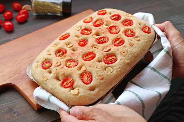Foto mão colocando uma panela de tomate italiano recém-assado e pão focaccia de alho na tábua de pão de madeira