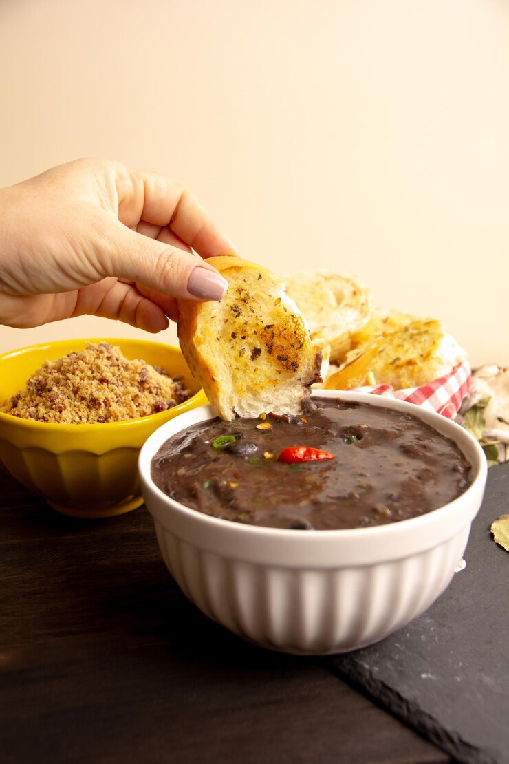 Foto mão com fatia de pão dentro de uma sopa cremosa de feijão preto com ervas frescas e pimenta