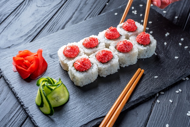 Mão com pauzinhos leva sushi de rolos de sushi seth com creme de queijo, arroz e salmão em uma placa preta decorada com gengibre e wassabi em uma mesa de madeira escura. comida japonesa