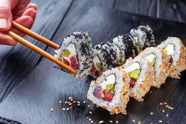 Mão com pauzinhos leva sushi de sushi rolls com creme de queijo, arroz e salmão no quadro negro decorado com gengibre e wassabi na mesa de madeira escura. Comida japonesa Foco suave seletivo