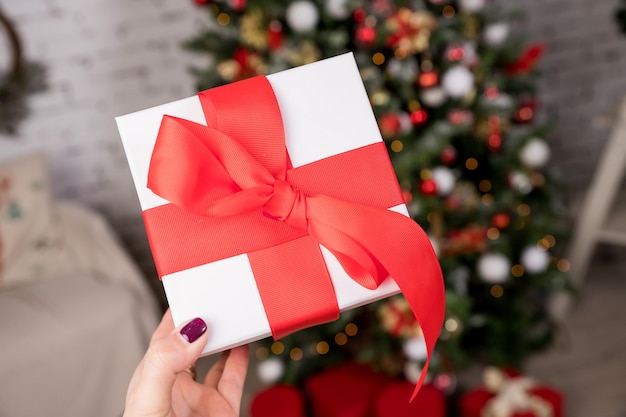 Mão com presente de Natal com fita vermelha perto da árvore na sala de estarUma árvore de Natal lindamente decorada com muitas caixas de presente de feriado bem embrulhadas conceito de clima aconchegante de férias