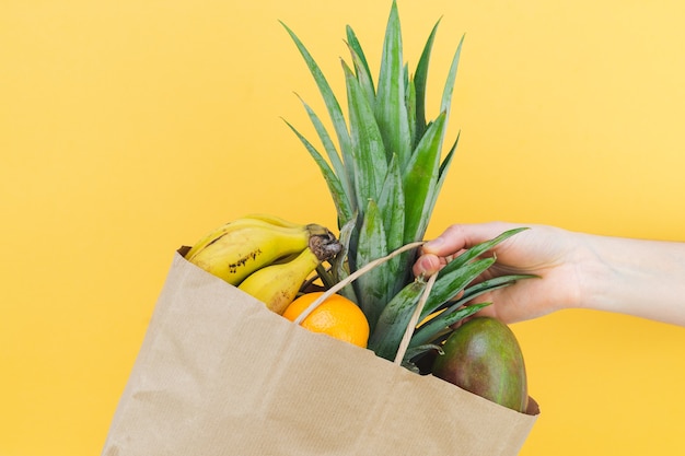 Mão com saco de papel com frutas sortidas em fundo amarelo.