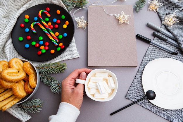 Mão com uma xícara de café, pirulitos, luzes de guirlanda de Natal, bloco de notas em branco e ramos de pinheiro
