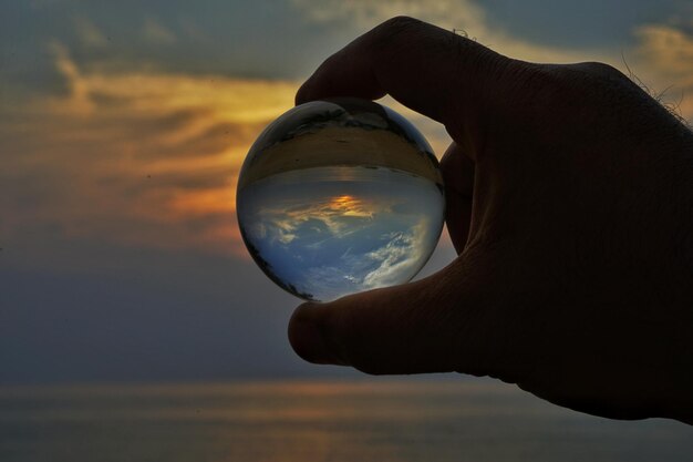 Mão cortada de pessoa segurando bola de cristal contra o mar e o céu durante o pôr do sol