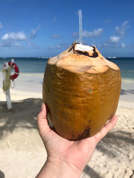 Foto mão cortada segurando coco na praia contra o céu azul