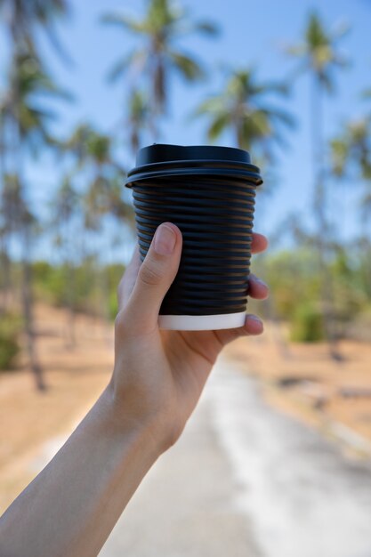 Mão da mulher com uma xícara de papel cartão de chá de café pelo parque com palmeira