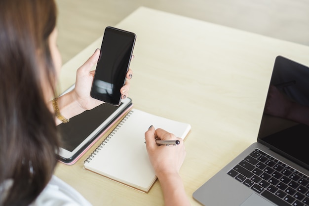 Mão da mulher que guarda o smartphone da tela vazia com portátil e a tabuleta digital na tabela.