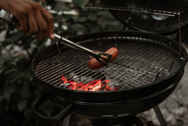 Mão de africano masculino com um garfo segurando a salsicha na churrasqueira com brasas. piquenique com amigos. lazer ao ar livre, celebração, alimentação pouco saudável.