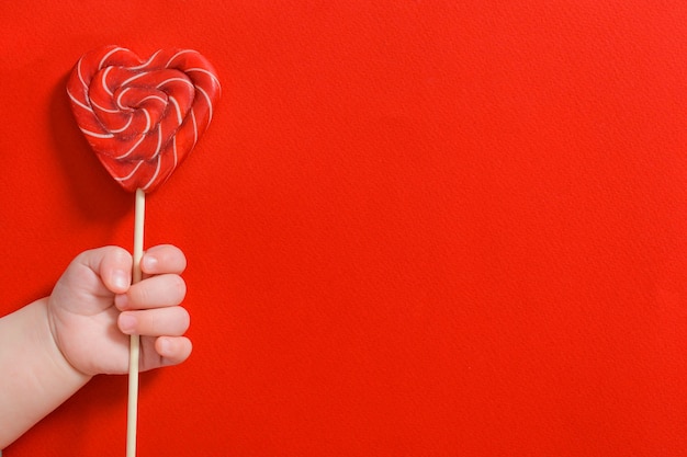 Mão de bebê recém-nascido segurando coração de doce no palito sobre fundo vermelho