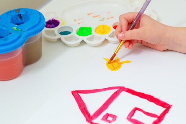 Mão de criança desenhando o sol amarelo com uma casa vermelha com pincel e aquarelas na folha de papel branca. Conceito de arte.