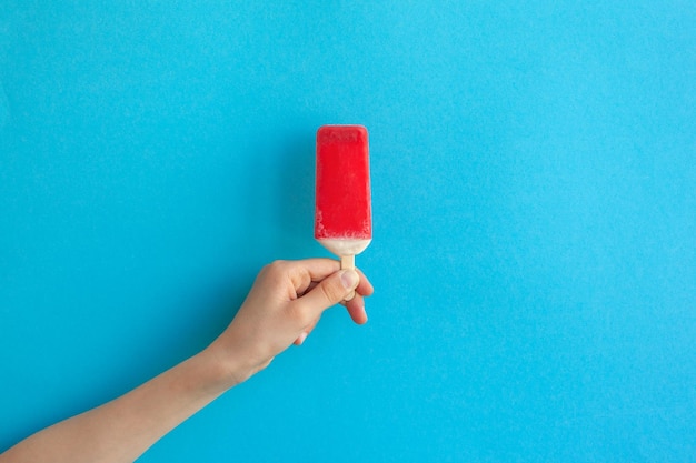 Mão de criança segurando picolé de sorvete de morango vermelho sobre fundo azul