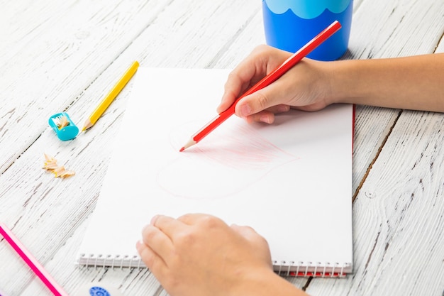 Mão de crianças desenhando coração vermelho com lápis de cor em papel branco na mesa de madeira