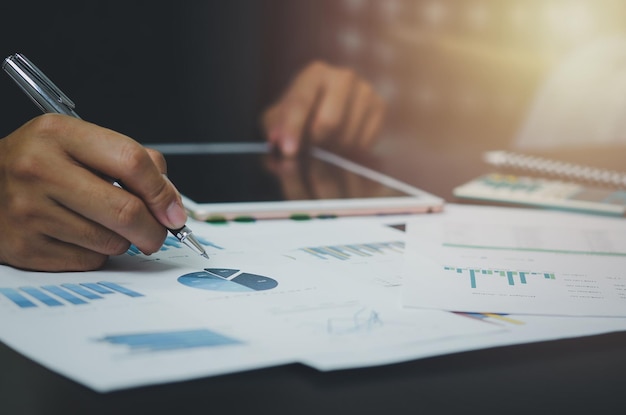 Mão de empresário segurando a caneta apontando a papelada gráfico relatório análise estatística e documento gráfico financeiro investimento marketing na mesa na empresa