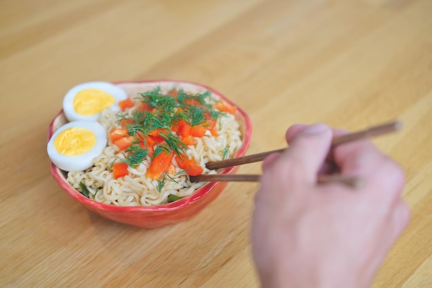 Mão de homem comendo macarrão de ovo asiático em tigela vermelha com pauzinhos de bambu close-up