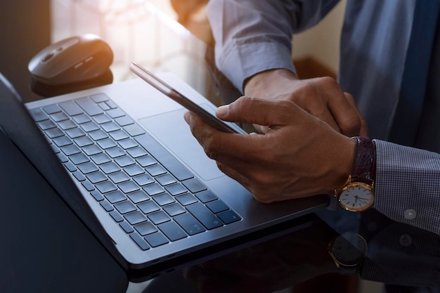 Mão de homem de negócios casuais usando telefone móvel inteligente e digitando no teclado do computador laptop com calculadora e papel milimetrado de dados na mesa do espelho.