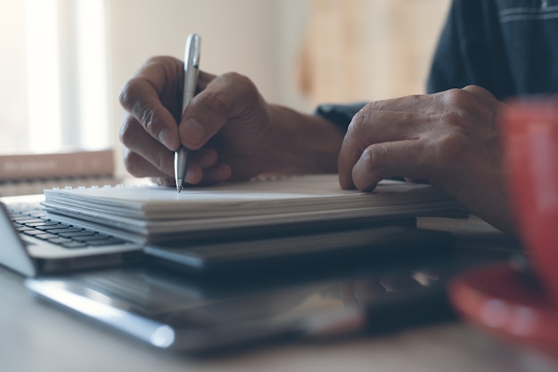 mão de homem escrevendo em um caderno de papel e trabalhando em um laptop no escritório