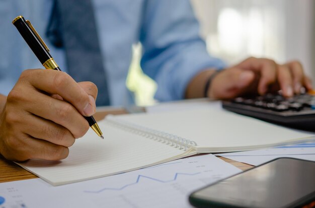 Mão de homem segurando caneta escrevendo relatório de gráfico de finanças de investimento de negócios de análise de livro na mesa