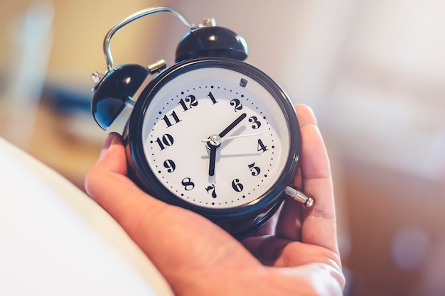 Mão de homem segurando o despertador para acordar às 7:00 da manhã, acorde na hora com efeito de luz no estilo vintage quente do quarto