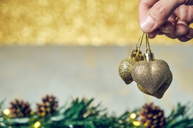 Mão de homem segurando um enfeite de corações dourados de natal com uma mesa de madeira de pinhas e um fundo dourado brilhante fora de foco