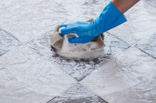 Mão de homem usando luvas de borracha azul está usando uma esponja para limpar o chão de ladrilhos.