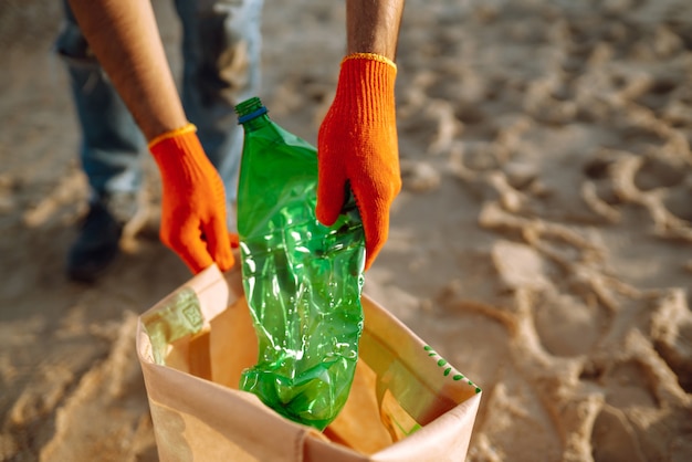 Mão de homens recolhe garrafa de plástico na praia do mar. o voluntário que usa luvas de proteção coleta o plástico da garrafa.