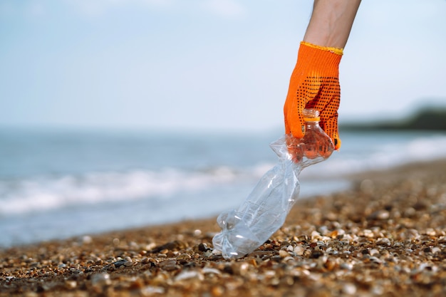 Mão de homens recolhe garrafa de plástico na praia do mar. O voluntário que usa luvas de proteção coleta o plástico da garrafa.