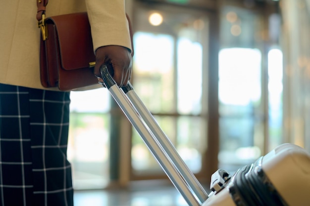 Mão de jovem negra em roupas casuais inteligentes com bolsa puxando mala