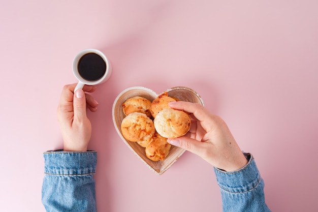 Mão de menina segurando pão de queijo brasileiro, vista de cima, fundo rosa, caneca de café, tigela em forma de coração