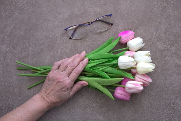 Mão de mulher adulta com buquê de tulipas e copos na mesa marrom, vista superior.