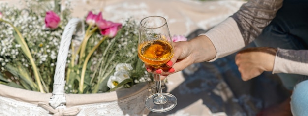 Mão de mulher closeup segurando um copo de vinho com vinho branco. Cesta com flores na manta de piquenique. Banner horizontal ou cabeçalho.