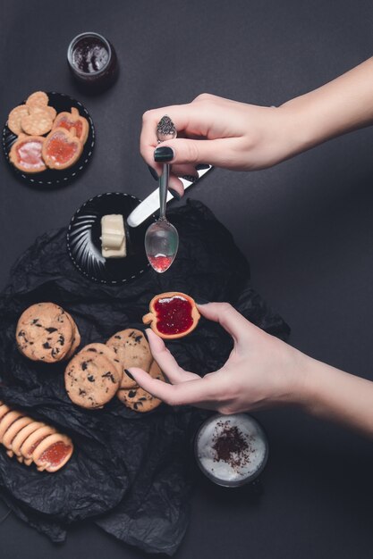 Mão de mulher com colher com geléia e biscoitos perto de xícara de café ou cappuccino e biscoitos de chocolate no fundo preto da mesa.