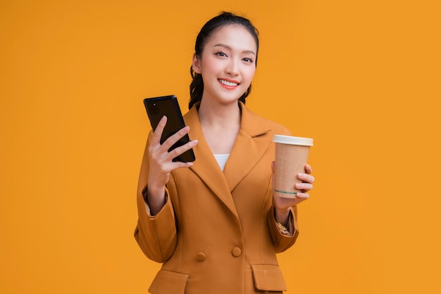 Mão de mulher de negócios asiática inteligente casual usando smartphone segure conversa de negócios de xícara de café sorrindo olhe para a câmera confiante expressão facial alegre estúdio tiro fundo amarelo