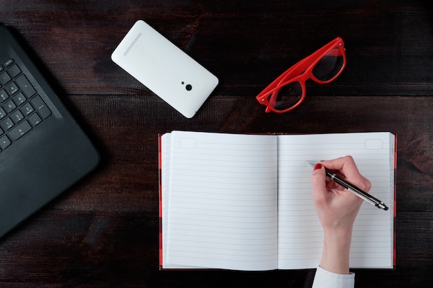 Mão de mulher escrevendo no caderno, conjunto de negócios de laptop óculos smartphone vista superior