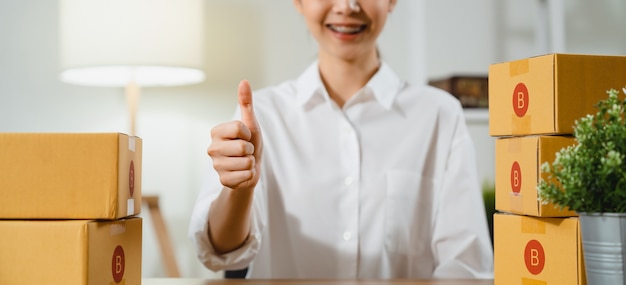 Mão de mulher fazendo o polegar para cima sinal e sorrindo e embalando produtos de entrega de caixa aos clientes na mesa.