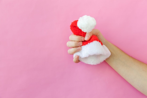 Mão de mulher feminina de design simplesmente mínimo segurando o chapéu de Papai Noel do enfeite de Natal isolado no fundo colorido pastel rosa na moda. Tempo de dezembro de ano novo de Natal para o conceito de celebração. Copie o espaço