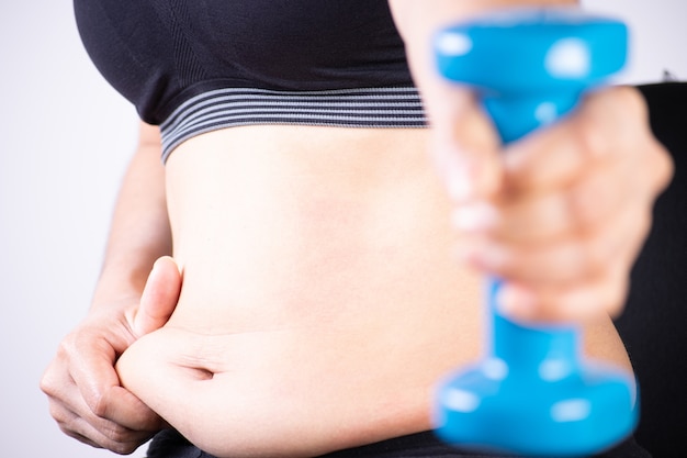 Foto mão de mulher gorda segurando halteres e tocando o excesso de gordura da barriga