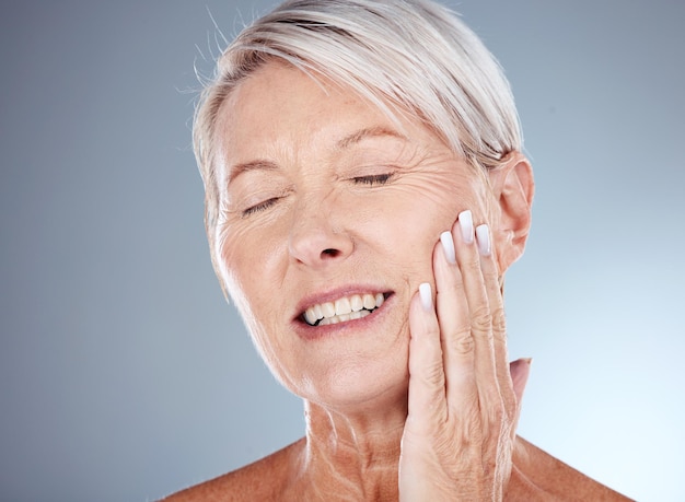 Mão de mulher idosa e sorriso para cosméticos para a pele ou hidratante contra um fundo de estúdio cinza Mulher sênior tocando rosto e sorrindo em satisfação para tratamento facial antienvelhecimento saudável