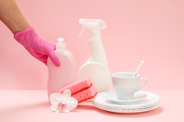 Mão de mulher na luva de nitrilo com garrafa de detergente e utensílio em um fundo rosa