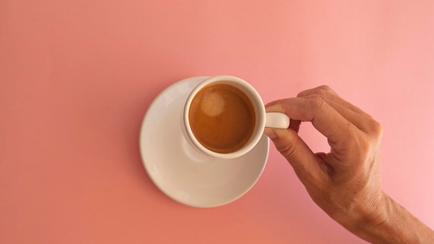 Mão de mulher pegando xícara branca com café em fundo rosa claro
