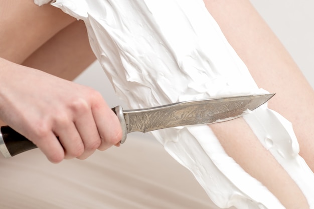 Mão de mulher raspa a perna por uma faca manchada com espuma de barbear no fundo branco. Conceito de depilação ou depilação.