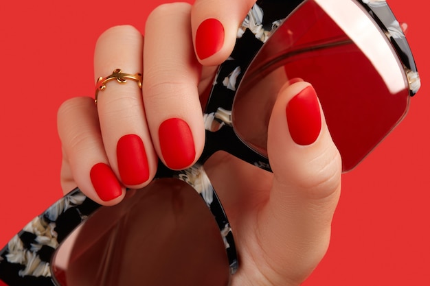 Mão de mulher segurando óculos de sol sobre fundo vermelho manicure e pedicure tendências de design