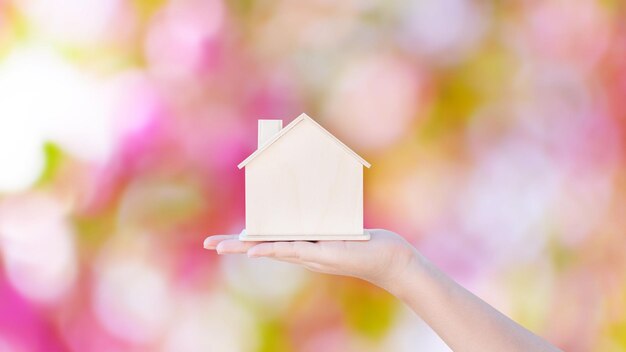 Mão de mulher segurando um pequeno modelo de casa de madeira com fundo abstrato de bokeh rosa vida familiar e conceito imobiliário de negócios casa ecológica