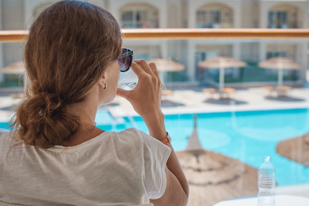 Mão de mulher segurar o copo de água na varanda do hotel contra a piscina