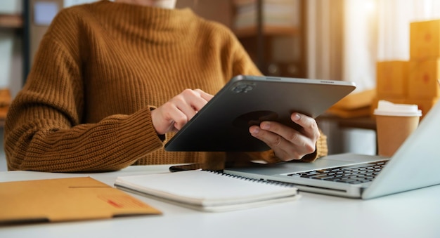Mão de mulher usando tablet e tablet e caderno de anotações no escritório de suas compras on-line de negócios Em casa xA