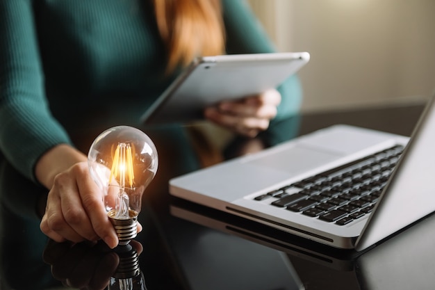 Foto mão de negócios segurando a lâmpada com o uso de computador portátil e a pilha de dinheiro no escritório. ideia de economia de energia e conceito de finanças contábeis à luz da manhã