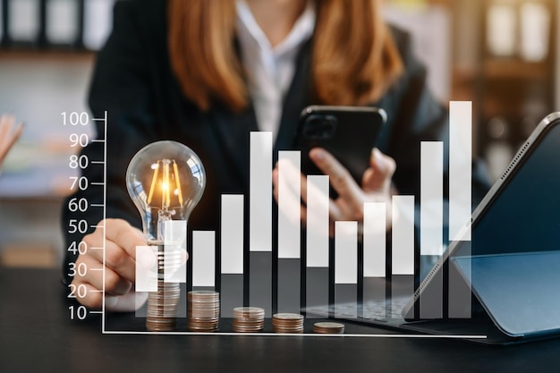 Mão de negócios segurando a lâmpada com o uso de computador portátil e pilha de dinheiro na ideia de escritório economizando energia e conceito de finanças contábeis na luz da manhã